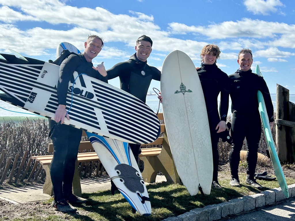 Four individuals are standing side by side in bright daylight, all wearing wetsuits, and holding different types of surfboards. They are smiling and seem to be in high spirits, likely either preparing for or returning from surfing. One person is giving a thumbs up to the camera. They are standing near what appears to be a coastal area with a clear blue sky in the background. There is a wooden fence partially visible behind them, and grasses can be seen at the bottom of the image. There's some text on one of the surfboards which appears to be a brand name.