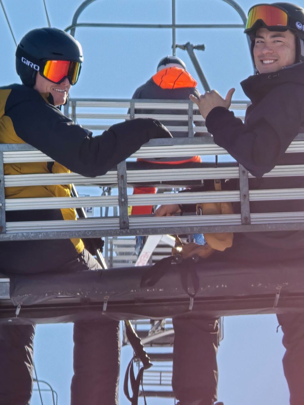 Two people are sitting on a chairlift, with both facing the camera and smiling. The person on the left is wearing a yellow and black jacket with a black helmet and reflective ski goggles. They are holding up their right fist as if knocking on an invisible door. The person on the right is dressed in a black jacket and also wearing a black helmet equipped with reflective ski goggles. They are making a 'hang loose' hand gesture with their right hand, which involves extending the thumb and little finger while holding the other fingers with the palm facing out. Behind them is a bright, clear sky, and the chairlift structure is visible above their heads. Although the image is presumed to be taken at a ski resort, no snow or specific ski equipment can be seen from this perspective.