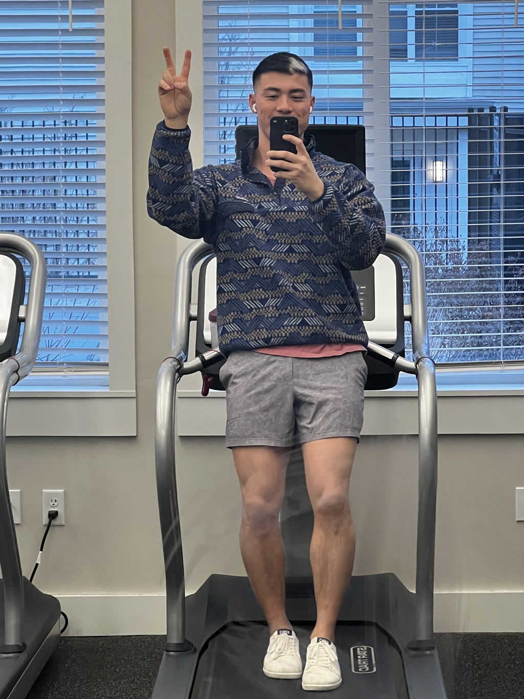 A person is standing on a treadmill, holding a phone in their right hand to take a mirror selfie. The individual is wearing a patterned long-sleeved sweater, gray shorts, and white sneakers. They are making a peace sign with their left hand raised next to their face. The background indicates an indoor setting with exercise equipment, a window with blinds, and the reflection of some furniture. The lighting is bright inside the room.