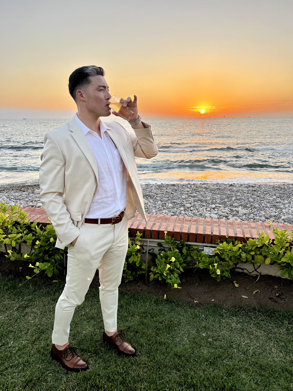 A person is standing on a grassy area in front of a beach during sunset, elegantly dressed in a cream-colored suit with a white shirt. They are holding a drink in a glass and appear to be sipping from it while enjoying the view. The ocean appears calm, and the sky is painted with hues of orange and blue as the sun sets on the horizon. Gentle waves can be seen touching the shoreline. There is a line of green shrubs behind the person and an unoccupied pebble border in the foreground.