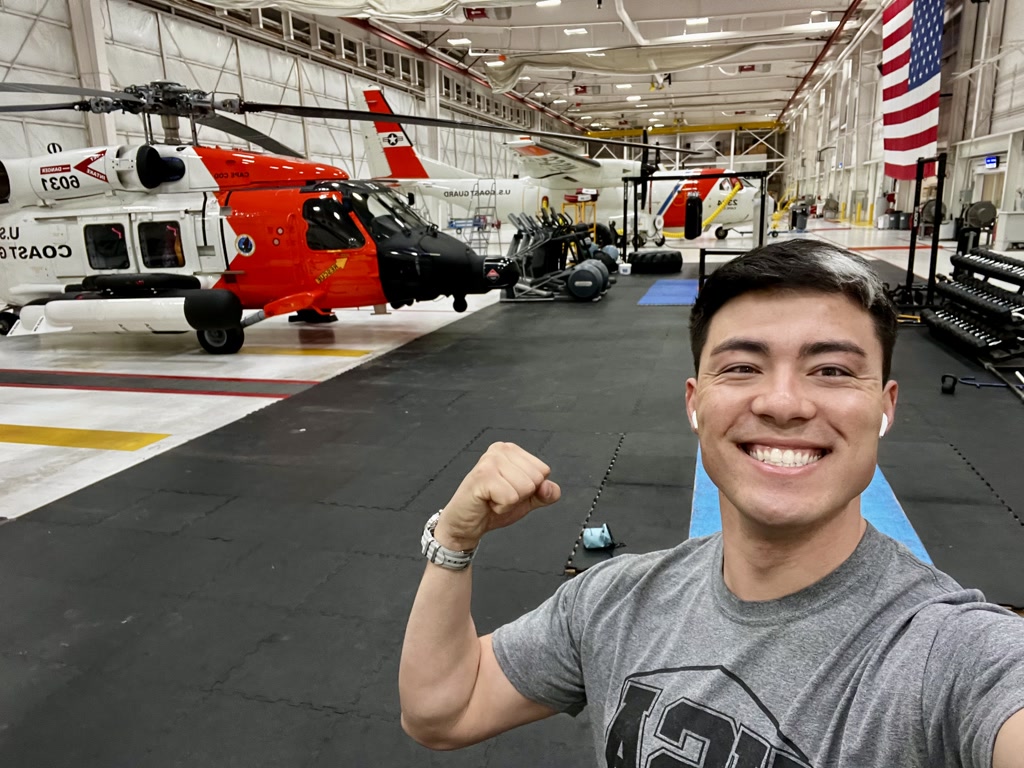 A person is smiling for a selfie with a flexed bicep pose in the foreground. Behind the individual, there is a large hangar-like space displaying several helicopters. Two of these helicopters are prominently featured; one is mainly white with red markings and a stylized American flag on the tail, and the other one is orange with white and black accents and also features a smaller American flag on the tail. On the far right, there is a gym setup with various weights and fitness equipment. The hangar has an industrial look with a high ceiling and bright lighting. The floor is marked with safety lines and there is a large American flag hanging from the back wall.