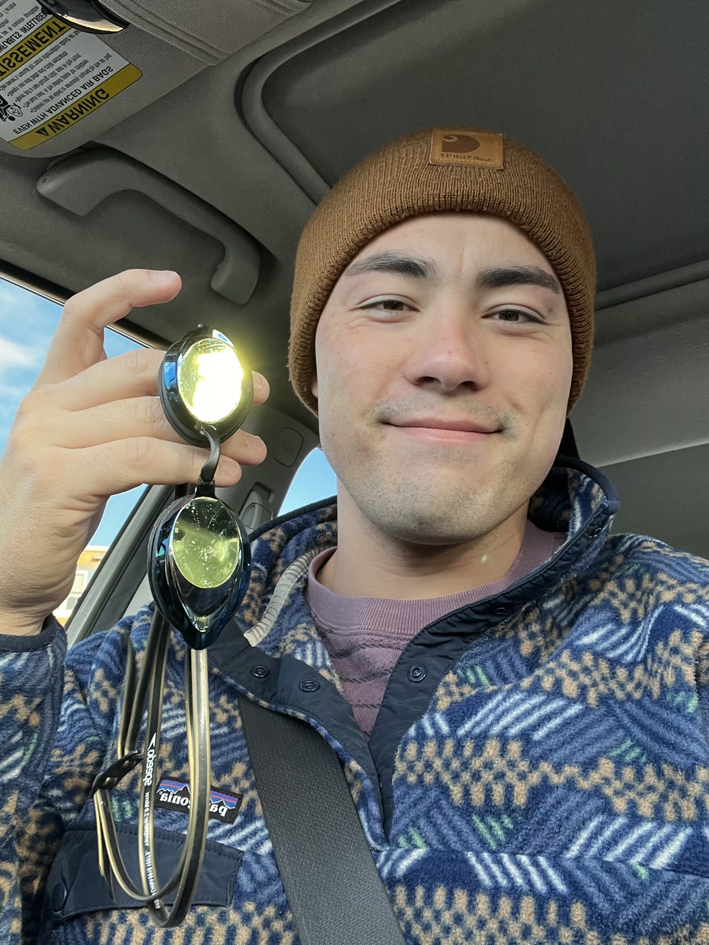 A person is sitting in the driver’s seat of a car, holding a stethoscope up in front of them with the diaphragm aimed towards the camera, causing it to reflect light. The person is wearing a brown beanie with a logo patch at the fold and a blue and white plaid fleece jacket over a maroon t-shirt. They appear to be smiling and wearing a seat belt. A sun visor with warning labels is visible in the background.