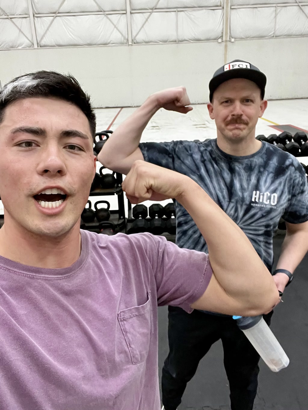 Two individuals, likely at a gym, are both flexing their arms to show off their muscles. The person in the foreground is seemingly taking a selfie with a smartphone; he is wearing a purple shirt with a small pocket and has a watch on his left wrist. The person in the background, wearing a black cap with a red and white logo and a dark tie-dye t-shirt with the letters 'H.CO' printed on it, is also flexing but with a more reserved expression. There's a rack of dumbbells visible behind them.