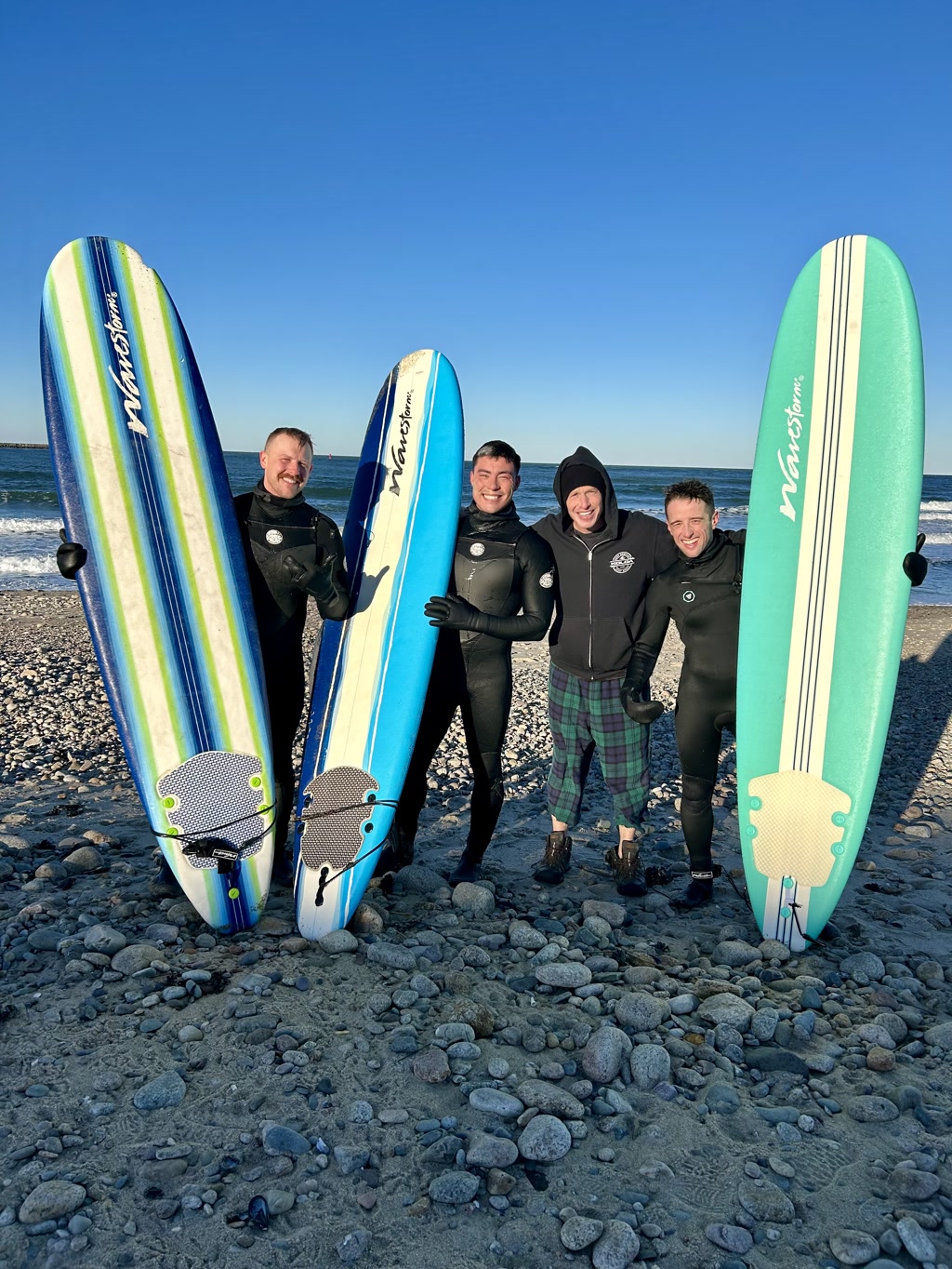 Four people are standing on a pebble-covered beach, each holding a surfboard vertically. They are wearing thick wetsuits, gloves, and booties, suggesting cold weather. The surfboards are colorful, ranging from blue to green shades, and appear to be made of sturdy foam, designed for buoyancy and stability. The sky is clear and blue indicating it's a sunny day, and the ocean is visible in the backdrop. The group is smiling and appears to be enjoying the moment. There is a mix of calmness and excitement evident in their postures.